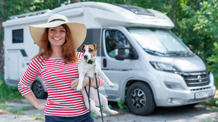 Caucasian red-haired woman in a straw hat and striped sweater walks with a small dog in front of a mobile home. Travel in a camper van with your pet