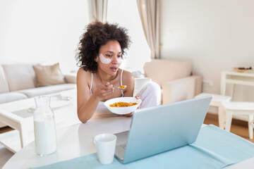 Obraz na płótnie Canvas Attractive woman using her laptop computer while having breakfast and studying in home indoors. Freelance African American woman working on her laptop in the morning . Cup of milk with corn flakes