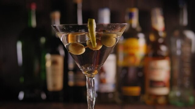 A glass of vermouth and gin with olives and lime on the bar against the background of blurred bottles, the glass rotates. Close-up.