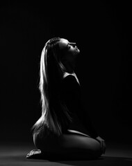 Black and white portrait of young sensual woman with long silky straight hair in dark bodysuit sitting side to camera on buttocks with eyes closed. Haircare, beauty, wellness, hairstyle concept