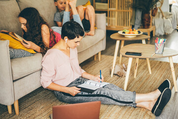 Fototapeta na wymiar Short haired woman with bluetooth earpiece writes in notebook sitting on floor near laptop while teenage girl and boy lies on sofa in light living room.