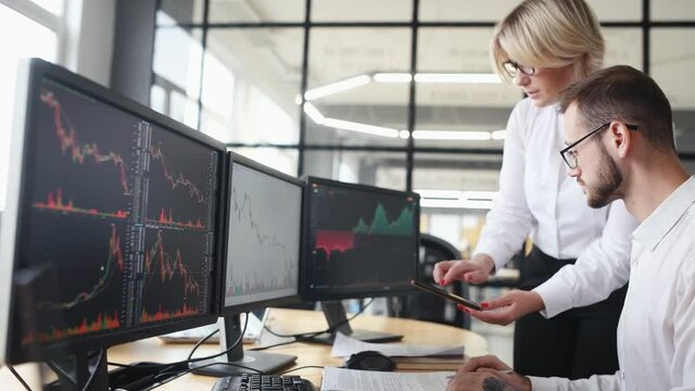 Woman walks in and showing information on smartphone and display. Male and female stockbrokers in formal clothes works in the office with financial market and graphs on monitors.