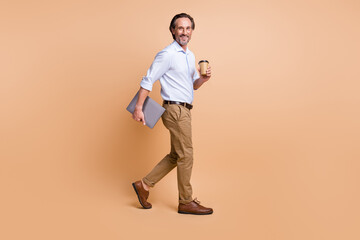 Fototapeta na wymiar Full length profile photo of guy hold coffee cup laptop walk wear specs shirt pants shoes isolated beige color background