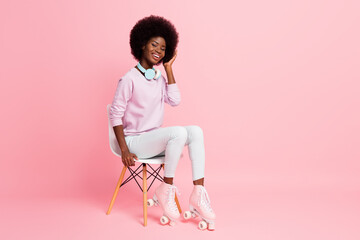 Portrait of pretty cheerful wavy-haired girl wearing roller skates sitting on chair isolated over...