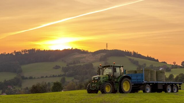 Timelapse close up of agricultural machinery tractor driving through a field and collecting bales of chopped hay on a hull during sunset over the horizon.