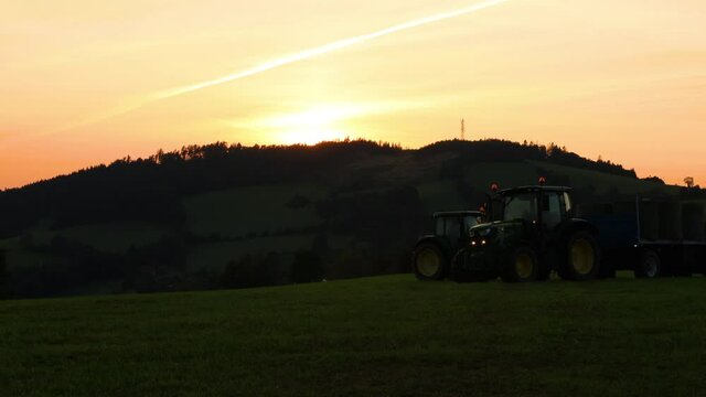 Timelapse of agricultural machinery tractor driving through a field and collecting bales of chopped hay on a hull during sunset over the horizon behing trees.