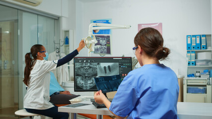 Stomatologist nurse comparing radiographics looking at computer, while specialist doctor with face mask speaks to man with toothache sitting on stomatological chair preparing tools for surgery
