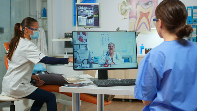 Assistent having video call with expert stomatologic dotor using computer while doctor is working with patient in background. Nurse listening dentist on webcam sitting on chair in stomatological