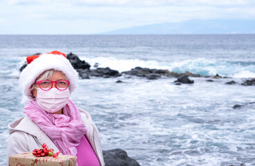 Obraz premium Senior woman in santa hat holding a christmas gift - wearing surgical mask due to coronavirus. Horizon over water