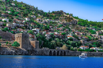 Fototapeta premium White boat in azure water against the background of an ancient arched wall and old town in Alanya (Turkey). Beautiful seascape with a historic resort city on the coast in the Turkish Riviera
