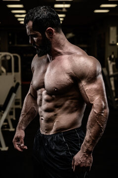 strong young bearded caucasian man with sport physique body torso standing in dark fitness gym with equipment