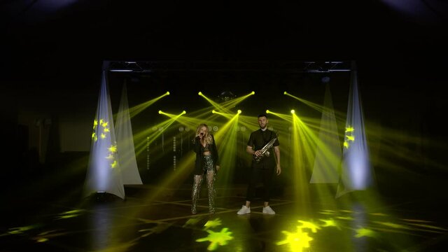 Musical band group of two people playing song, performing on concert musician stage with lights. Shooting a music video. Singer vocalist girl, saxophonist sax man. Actors dancing, singing. Slow motion