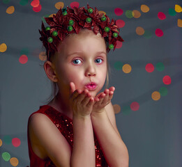 Obraz na płótnie Canvas Christmas kiss. Kid girl wearing red sequin dress and Christmas wreath. Blond hair and blue eyes posing and fool around in studio on grey background with illumination and boke lights.