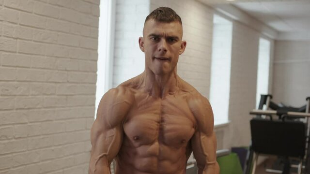 A young bodybuilder with a naked torso stands in the gym and bother his muscles by clenching his teeth and posing for the camera