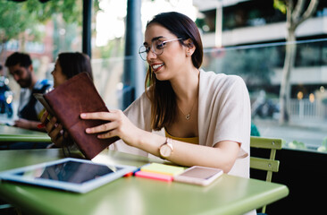 Happy female journalist checking informative notes in education textbook sitting in street cafe, successful hipster girl in optical eyewear for vision correction using personal planner outdoors