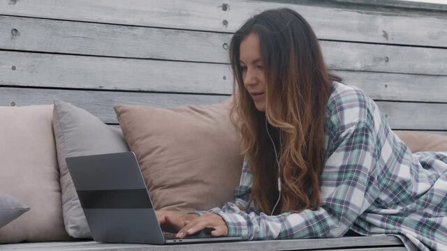 Medium shot of young attractive woman with long hair wearing casual clothes lying on wooden bench on terrace and typing on laptop keyboard