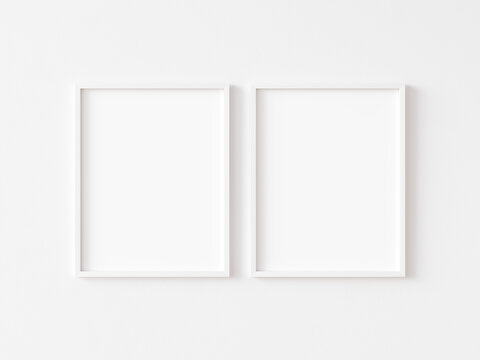 Two blank vertically oriented rectangular picture frames with thin white border hanging on white wall. 3D illustration.