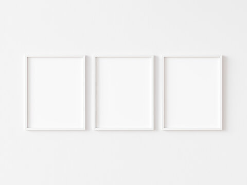 Three blank vertically oriented rectangular picture frames with thin white border hanging on white wall. 3D illustration.