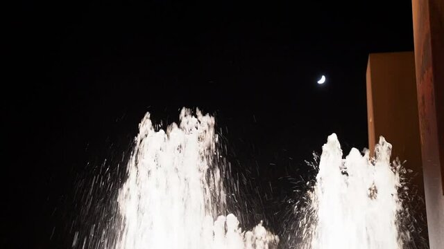 Low angle shot of white splashing water streams of fountain against black night sky with crescent half moon