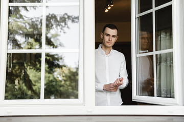 Young man in white shirt buttoning a button at home. Groom's gathering. groom at home. the guy gets dressed. the man on self-isolation looks through the window