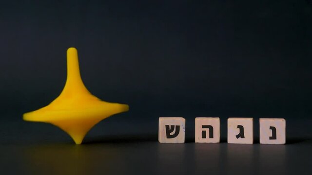 A plastic Hanukkah spinning top spins beside the four hebrew letters nun, gimel, heh and shin shown against a black background.