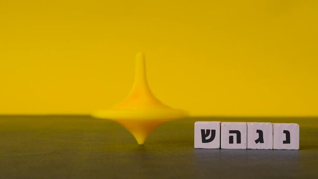 A plastic top spins and wobbles to a stop beside the four hebrew letters nun, gimel, heh and shin shown against a two-tone background.