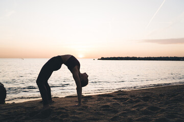 Flexible female yogi training Urdhva Dhanurasana during evening workout at beach seashore, fit girl training concentration and body balance for keeping healthy lifestyle and wellbeing vitality