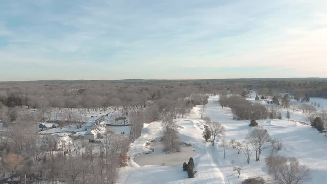 Level drone view of a snowy golf course abutting a neighborhood and a cemetery. Small pond comes into view and tracks, trees and snow visible below.