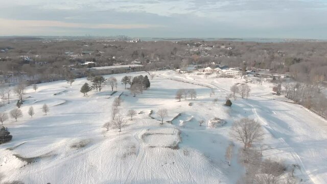 A leftward pan over a snowy golf course to reveal Boston, MA, USA on the horizon. Drone view with water and other features in the distance.