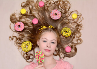 A beautiful young fashion model girl holds a multi-colored lollipop in her hands and lies on a background of pink and yellow donuts and macarons. Sweet life concept. Junk food