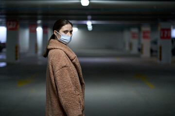 Woman in a medicine mask and beige coat in an underground parking lot in a winter day, protection against virus and infections during an epidemic.