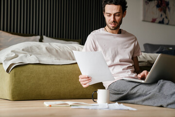 Focused man working with papers and laptop while sitting on floor