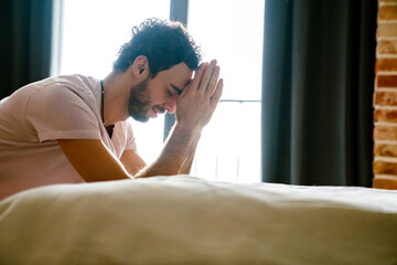 Unhappy unshaven man praying while holding hands together in bedroom