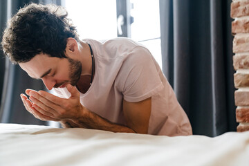 Focused unshaven man praying while holding hands together in bedroom