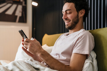 Joyful brunette man smiling and using mobile phone while lying in bed