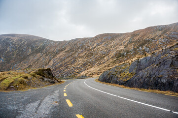 Dark wet road near sun lit cliffs going from left to right in Ireland near Atlantic ocean in a rainy day in winter and autumn. Concepts: season, outdoors, travel, landscape