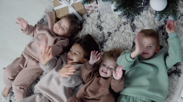 Four sweet children lay down jn a carpet near Christmas tree and waving their legs and hands. View from top