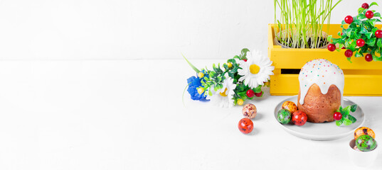 Fototapeta premium Easter composition on the white table. Easter cake with sugar icing, colored candy eggs, flowers and sprouted grass in the yellow box. Copy space. Wide banner