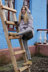 A joyful attractive blonde girl, in warm clothes, stands on a wooden staircase near the house.