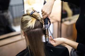 Naklejka premium Young woman hairdresser making curls at blond hair with curling irons at luxury barber salon. Stylist and beauty