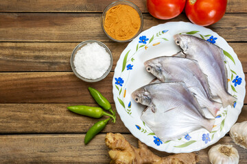 Fresh white pomfret fish in a plate along with ginger, garlic, green chilli and spices on wood background.
