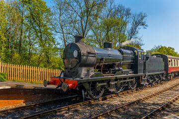 A steam train approaching a station on a railway line in Sussex, UK on a sunny summer day