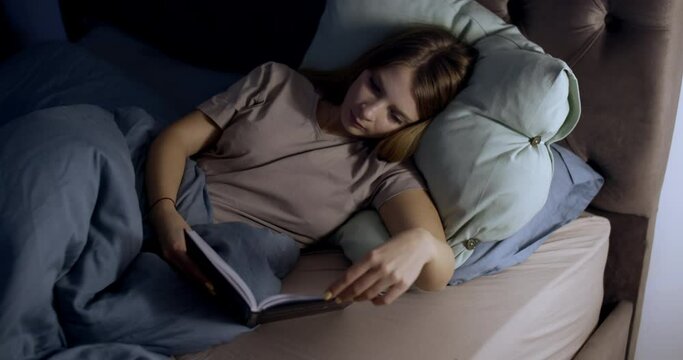 a young woman reads a book in the evening in the bedroom bed, slow motion.
