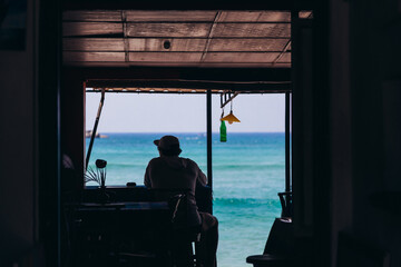 A silhouette of a man, sitting in a vintage style cafe on a beach in front of blue ocean panorama, enjoying vacation moment. Travel relax concept background.