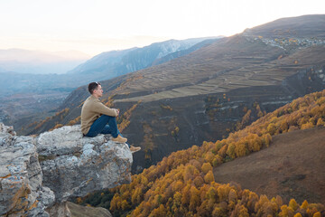 Obraz premium Guy in brown shirt and blue jeans sits on large hilltop rock edge against orange yellow forests and fields of mountainous landscape