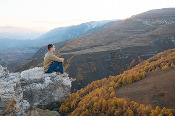 Guy in brown shirt and blue jeans sits on large hilltop rock edge against orange yellow forests and fields of mountainous landscape