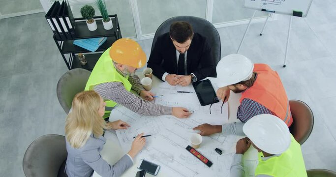 Top view of likable confident professional diverse team of engineers in vests and hardhats which brainstorming with their male and female managers over building project in meeting room