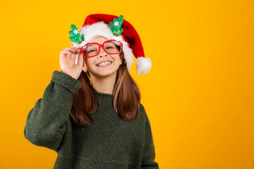 Portrait of young girl wearing traditional Santa Claus hat and green knitted sweater posing over isolated yellow background. Studio shot of a kid celebrating Christmas. Close up, copy space.