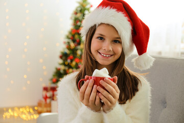 Young girl wearing white fluffy sweater holding a cup of cacao with bunch of marshmallows at home. Christmas tree with stack of presents underneath, festive bokeh light. Blurry background, copy space