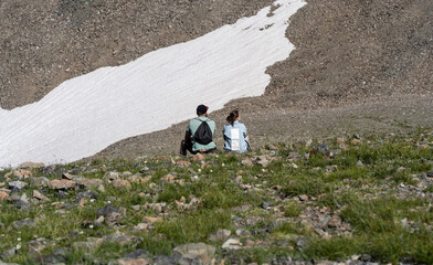 Rear view of man and woman sitting on rock and resting in mountains. Married couple enjoy beautiful view of mighty mountains in sunny weather.